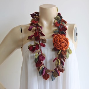May include: A crocheted necklace with a large orange flower and leaves in shades of green, red, and brown.