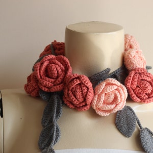 May include: A handcrafted crochet floral necklace featuring coral and peach-colored roses with gray leaves and stems. The necklace is displayed on a mannequin, showcasing the intricate details of the handmade accessory. A unique, wearable art piece.