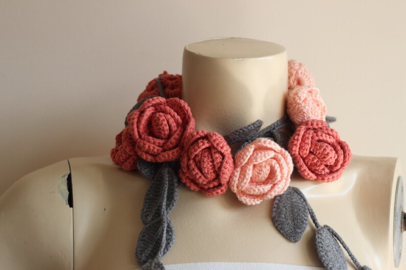 May include: A handcrafted crochet floral necklace featuring coral and peach-colored roses with gray leaves and stems. The necklace is displayed on a mannequin, showcasing the intricate details of the handmade accessory. A unique, wearable art piece.