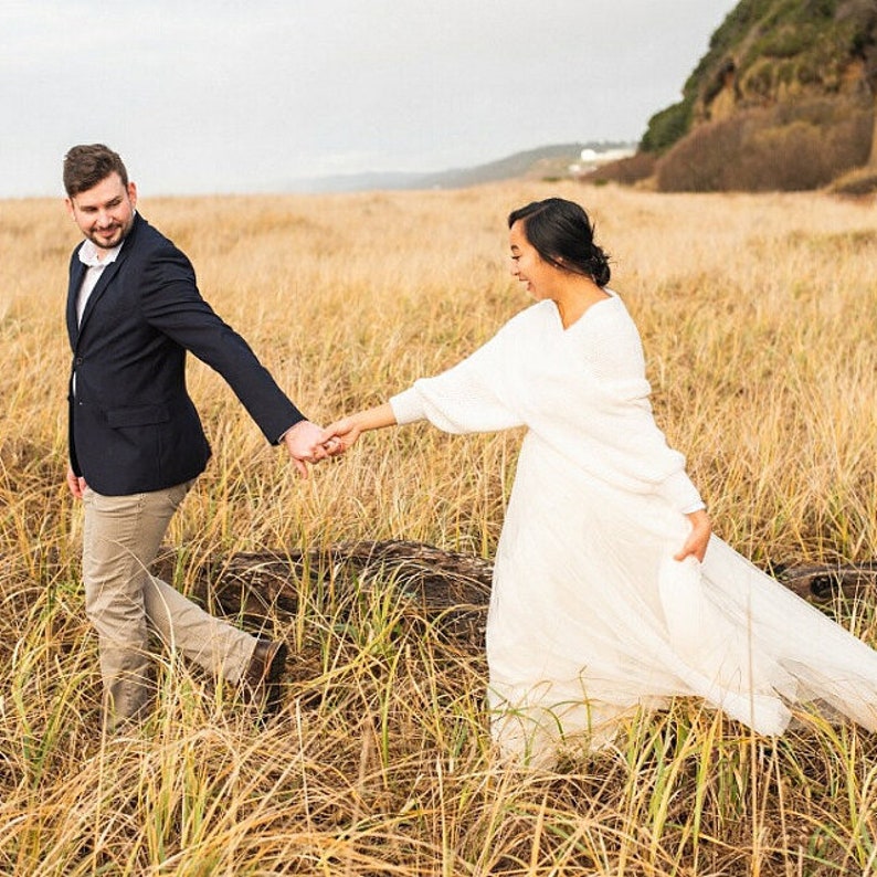 May include: A couple walks hand-in-hand through a field of tall, dry grass. The woman is wearing a white dress and the man is wearing a navy blue suit jacket and khaki pants.