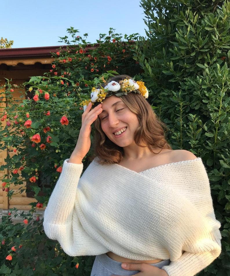 May include: A woman wearing a white off-the-shoulder sweater with a flower crown on her head. The crown is made of white flowers and yellow leaves.