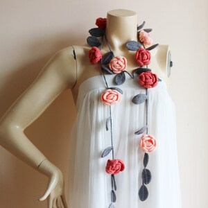May include: A handmade crochet floral garland featuring red and peach roses with gray leaves. The garland is draped on a mannequin wearing a white dress. The roses are evenly spaced along a dark gray stem.