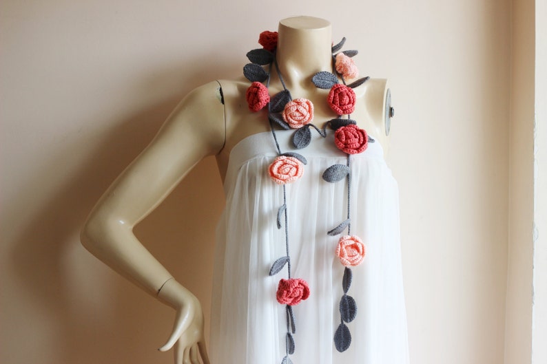 May include: A handmade crochet floral garland featuring red and peach roses with gray leaves. The garland is draped on a mannequin wearing a white dress. The roses are evenly spaced along a dark gray stem.