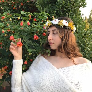 May include: A woman wearing a white knitted sweater and a flower crown with white and yellow flowers. The crown is made of fabric flowers and greenery.