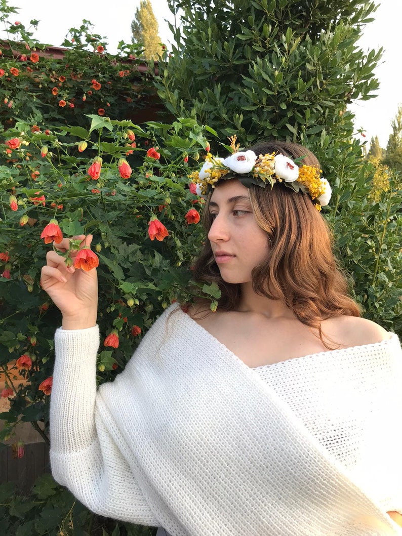May include: A woman wearing a white knitted sweater and a flower crown with white and yellow flowers. The crown is made of fabric flowers and greenery.