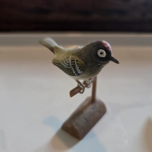 Op de afbeelding: Een gedetailleerd beeldje van een kleine vogel die op een houten tak zit. De vogel heeft een donkergrijze kop met een rode vlek, witte oogringen en een geel en grijs lichaam. Het beeldje staat tegen een witte achtergrond.