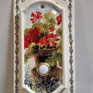 May include: Illuminated doorbell plaque with a floral design. The rectangular ceramic plaque features a watercolor painting of red geraniums in a brown pot, with a white button and gold trim. The text "ILLUMINATED DOORBELL PLAQUE" is at the top.