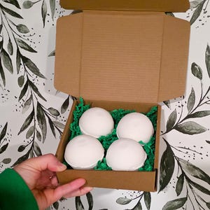 Op de afbeelding: Een bruine kartonnen doos gevuld met vier witte, bolvormige badbruisballen op groen papier. De doos wordt vastgehouden door een persoon met een groene mouw tegen een bloemenbehang.