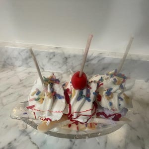 May include: A banana split dessert with three scoops of vanilla ice cream, topped with colorful sprinkles, red cherry, and drizzled with red syrup. The dessert is served in a clear, oval-shaped dish.