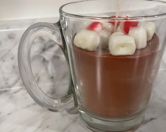 Puede incluir: Una vela en forma de taza de vidrio transparente llena de cera marrón, cubierta con trozos de cera blancos y rojos que parecen malvaviscos. Una sola mecha está centrada en la vela. La taza tiene un asa transparente.