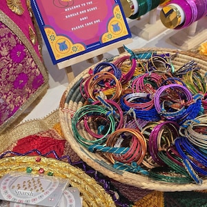 May include: A vibrant display of bangles, bindis, and decorative items. A sign reads "Bangle Bindi Bar" with the text "Welcome to the Bangle and Bindi Bar. Please take one." The scene includes a variety of bangles in a woven basket.