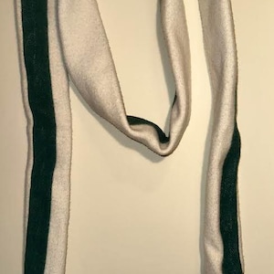 Könnte beinhalten: Cremefarbener Schal mit dunkelgrünem Mittelstreifen. Er hat an einem Ende einen braunen Lederaufnäher und am anderen eine kleine braune Schlaufe. Der Schal ist aus einem weichen, strukturierten Material gefertigt.