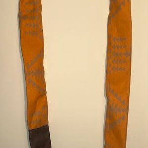 Op de afbeelding: Een oranje camerariem met een grijs geometrisch patroon. De riem heeft een bruin leren uiteinde en een zwart koord voor bevestiging. De riem is ontworpen om om de nek te worden gedragen.