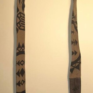 Könnte beinhalten: Ein langer Schal mit einem braunen und schwarzen geometrischen Muster. Der Schal hat ein dunkelbraunes, lederartiges Endstück. Der Schal hängt vor einem neutralen Hintergrund.