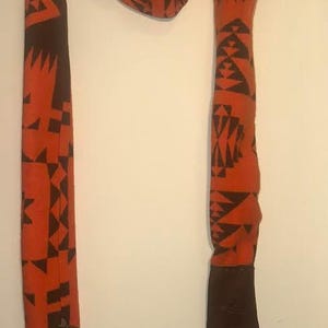 Op de afbeelding: Een oranje en zwart patroon sjaal met een geometrisch ontwerp. De sjaal is gemaakt van een zacht materiaal en heeft aan het uiteinde een donkerbruin lederachtig zakje. Een donkerbruin leren koord is aan de sjaal bevestigd.