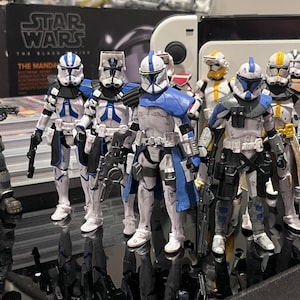 Puede incluir: Una colección de figuras de acción de Star Wars, incluyendo varios Clone Troopers y otros personajes. Las figuras son predominantemente blancas, azules y grises, con armaduras y armas detalladas. Las figuras están sobre una superficie reflectante.