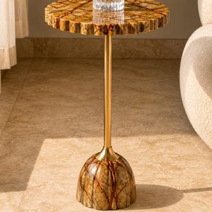 May include: A side table featuring a round, scalloped-edge top and a dome-shaped base, both crafted from brown and tan veined stone. A slender, polished gold-tone metal rod connects the base to the top, which holds a clear glass tumbler.