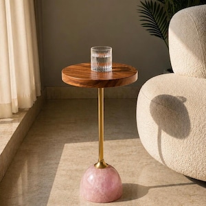 Puede incluir: Una mesa auxiliar con un tablero circular de madera oscura, un vástago fino de metal dorado y una base redondeada de cuarzo rosa pulido. Un vaso transparente con estrías verticales lleno de agua se sitúa en el centro de la superficie.