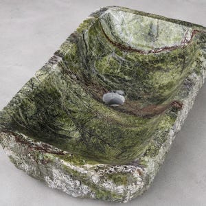 Puede incluir: Un lavabo rectangular de piedra tallado en mármol verde con vetas oscuras y un cuenco interior pulido. El exterior presenta una textura de roca natural y rugosa con depósitos minerales blancos y grises, y una abertura de drenaje circular central.