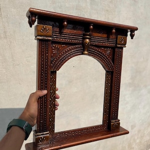 May include: A dark brown wooden wall shelf featuring an arched opening, intricate carved detailing, and small decorative finials along the top edge. The frame displays vertical textured patterns and gold-toned accents on the corners and center.