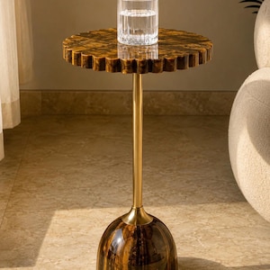May include: A side table featuring a circular, scalloped-edge top and a dome-shaped base, both crafted from brown and gold tiger's eye stone. A slender, polished gold-tone metal rod connects the base to the top, which holds a clear glass tumbler.