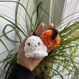 May include: A pair of handmade crochet Halloween decorations. One is a white ghost with black eyes, and the other is an orange pumpkin with a green stem. Both are held in a hand, with green plants in the background.