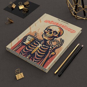 May include: A journal with a skeleton illustration holding a coffee cup that says "TRCK OR DRINK". The journal cover reads "UNDIAGNOSED" and "My Journal". The journal is beige, red, and black.