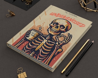 May include: A journal with a skeleton illustration holding a coffee cup that says "TRCK OR DRINK". The journal cover reads "UNDIAGNOSED" and "My Journal". The journal is beige, red, and black.