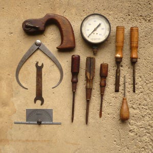 May include: A collection of vintage tools including a wooden-handled wheel tool, a metal caliper, a small wrench, a circular pressure gauge, a metal protractor, and several screwdrivers and chisels with wooden handles arranged on a textured surface.