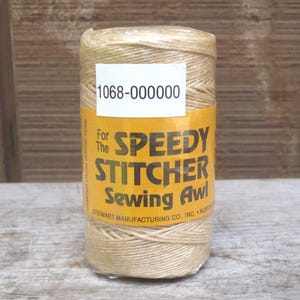 May include: A cylindrical spool of beige thread for a Speedy Stitcher Sewing Awl. The label is yellow with black text, including the product name and the text "Unwind from inside tube". A white sticker with the number "1068-000000" is also visible.