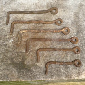 May include: A set of six hand-forged iron gate latches, each featuring a twisted metal shank, a circular eyelet at one end, and a right-angle hook at the other. The metal components exhibit a weathered, rusted brown patina throughout the surface.