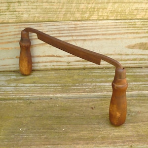 May include: A drawknife featuring a long, straight, rusted metal blade positioned horizontally between two vertical, cylindrical wooden handles. The tool has a weathered appearance with visible oxidation on the steel and a worn finish on the wood.