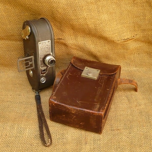 May include: A vintage dark brown Keystone movie camera with a metal viewfinder, a circular lens, and a black wrist strap, positioned next to a rectangular brown leather carrying case featuring a silver-toned metal latch closure mechanism.
