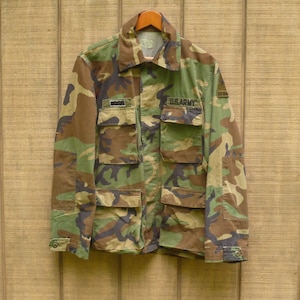 May include: A long-sleeve camouflage field jacket featuring a woodland pattern of green, brown, and black. The jacket includes a pointed collar, four front pockets with buttoned flaps, and black embroidered patches reading U.S. ARMY and a name tag.