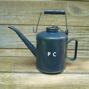 May include: A dark blue metal oil can featuring a long, curved spout, a cylindrical body with the letters P C printed in white, a side handle, and a top handle with a matching blue grip. A small metal chain connects the cap to the top handle.