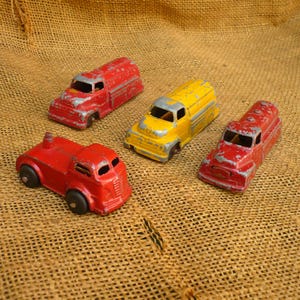 May include: Four vintage die-cast metal toy trucks including two red tanker trucks, one yellow tanker truck, and one red truck with a small chimney-like feature on the back. Each vehicle shows signs of paint wear and has a weathered appearance.