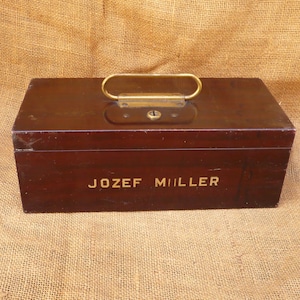 May include: A rectangular, dark brown wooden box with a brass handle and keyhole. The name "JOZEF MILLER" is printed in gold lettering on the front of the box. The box is sitting on a burlap surface.