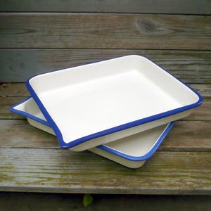 May include: Two white enamelware baking dishes with a blue trim are stacked on a wooden surface. The dishes have a square shape with slightly curved edges. The blue trim provides a contrasting border to the white enamel.