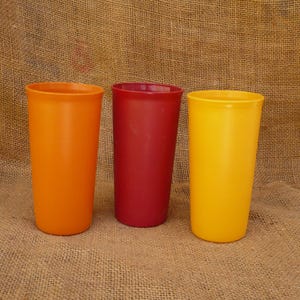 May include: Three tall, cylindrical plastic tumblers in vibrant colors: orange, red, and yellow. The tumblers are evenly spaced and stand upright against a textured, neutral-toned background. These are ideal for serving cold beverages.