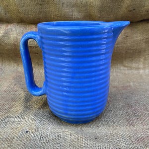 May include: A bright blue ceramic pitcher featuring a series of horizontal, raised ridges encircling the entire body. The vessel includes a curved handle on the left side and a prominent, pointed pouring spout on the upper right rim.