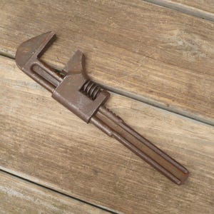 May include: An antique adjustable wrench with a rusty brown finish. The wrench features a movable jaw and a threaded adjustment mechanism. The tool is designed for gripping and turning nuts and bolts.