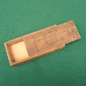 May include: A vintage wooden box with a sliding lid, featuring the text "LUFKIN MICROMETER" and "THE LUFKIN RULE CO." in black lettering. The box is open, revealing an empty interior. The box is made of light brown wood.