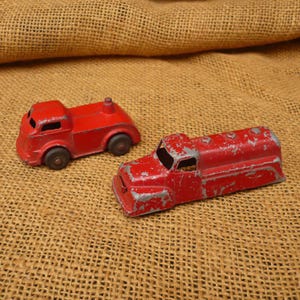 May include: Two vintage red toy trucks with black wheels. One truck has a rounded cab and a small smokestack. The other is a tanker truck with a rectangular cab. Both show signs of wear and tear.