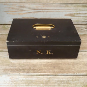 May include: A black wooden box with a gold-colored handle and the initials "N. K." in gold lettering. The box has a rectangular shape and is sitting on a light-colored wooden surface.