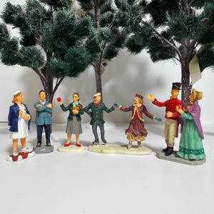 May include: A collection of miniature figurines depicting various characters, including a nurse, a man in a blue coat, a woman holding an apple, and others in winter attire. The figurines are set against a backdrop of artificial evergreen trees.