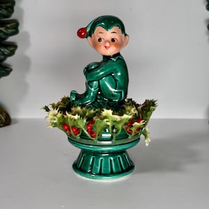 May include: A vintage ceramic Christmas elf figurine. The elf is dressed in a glossy green outfit with a red pom-pom hat. It sits atop a pedestal surrounded by holly and red berries. The base is a matching green color.