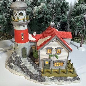 May include: A decorative ceramic lighthouse and house figurine with a red and white lighthouse, a white house with a red roof, and a small green door. The figurine has a snowy base and is surrounded by miniature evergreen trees.