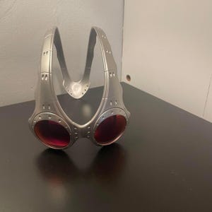May include: Silver-colored sports goggles with red lenses. The goggles have a unique, angular design with a bridge and side arms. The lenses are oval-shaped and set in a metallic frame.