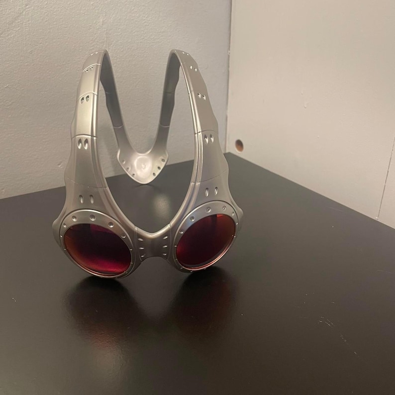 May include: Silver-colored sports goggles with red lenses. The goggles have a unique, angular design with a bridge and side arms. The lenses are oval-shaped and set in a metallic frame.