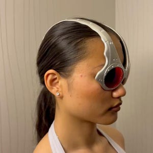 May include: Silver and red goggles with a curved, metallic frame and round lenses. The goggles have a silver headband and are worn on the head. The lenses are a deep red color.